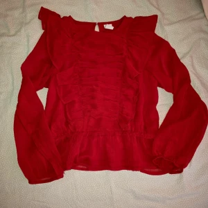 Röd volangblus - Säljer en röd blus med volangdetaljer framtill och långa ärmar. Blusen har en rund halsringning och knäppning bak i nacken. Perfekt för dig som vill ha något färgstarkt och trendigt.