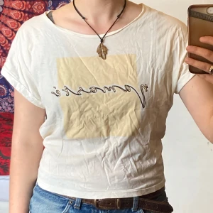 Vit t-shirt med tryck från Lindex, M - Vit t-shirt från Lindex med korta ärmar och ett beige fyrkantigt tryck framtill med texten 'Namasté'. Skön passform och tillverkad i mjuk bomull. Säljer då den känns för lite för mig. Nyskick.