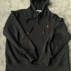 Svart hoodie från Ami Paris - Svart hoodie från Ami Paris med broderat rött hjärta och A på bröstet. Tröjan har huva med snörning, känguruficka och ribbade muddar. Tillverkad i mjuk bomull, perfekt för en chill och stilren look.