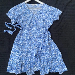 Blåvit blommig omlottklänning - Säljer en söt blåvit omlottklänning med blommigt mönster och korta ärmar. Klänningen har volangdetaljer nertill och knyts i midjan för en fin passform. Perfekt för varma dagar!