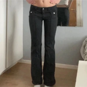 Svarta bootcut byxor med fickor - Snygga svarta byxor med bootcut-modell och markerade sömmar framtill. Byxorna har låg midja, två framfickor med silverfärgade knappar och är tillverkade i ett stretchigt material som sitter skönt. Perfekta för en trendig och avslappnad look.