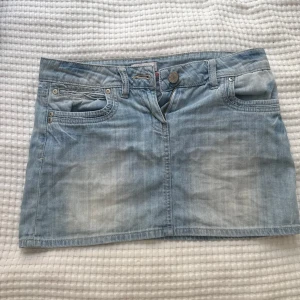 Ljusblå jeanskjol med fickor - Säljer en ljusblå jeanskjol med klassiska fem fickor, dragkedja och knapp framtill. Kjolen har bälteshällor och en liten dragkedjedetalj på ena bakfickan. Perfekt för en avslappnad stil.
