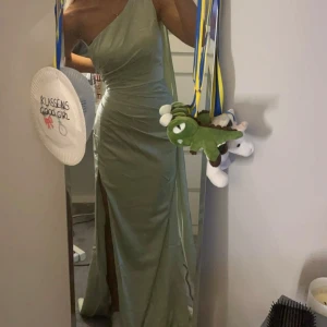 Balklänning - Säljer en elegant ljusgrön långklänning med oneshoulder och hög slits framtill. Klänningen har en figurnära passform och är perfekt för bal eller fes eller bröllop. . Materialet har en lätt glansig yta som ger en lyxig känsla. Klänning är skräddar sydd i midja och byst.