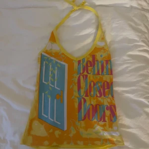Gul halterneck-linnetopp med tryck - Säljer en färgglad gul halterneck-linnetopp med tryck i blått, rött och vitt. Toppen har texten 'Behind Closed Doors' och en dörr-illustration framtill. Knytning i nacken och stretchigt material. Perfekt för sommarens alla tillfällen!