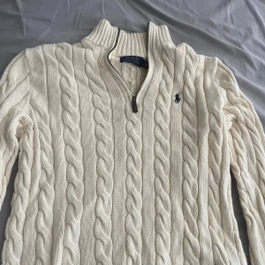 Vit kabelstickad tröja med halv dragkedja från Polo Ralph Lauren - Säljer en klassisk vit kabelstickad tröja från Polo Ralph Lauren med halv dragkedja och hög krage. Tröjan har lång ärm och den ikoniska broderade loggan på bröstet. Perfekt för en stilren och tidlös look.