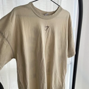 Beige oversized t-shirt från Gymshark - Säljer en beige t-shirt från Gymshark med stort tryck på ryggen och liten logga framtill. T-shirten har en oversized passform. Fick den i julklapp men den är för stor för mig