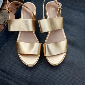 Guldiga sandaler med klack från H&M stl 41 - Säljer ett par snygga sandaler i metallic guld från H&M. Skorna har breda remmar, öppen tå och en stadig klack. Perfekta för dig som vill sticka ut med ett par trendiga och bekväma skor.