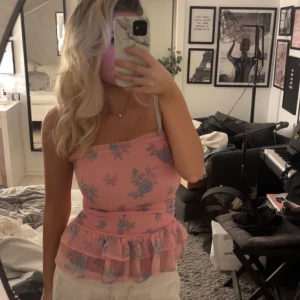 Rosa blommig tube topp - Söt rosa topp med blått blommönster och volang nertill. Axelbandsmodell med figurnära passform och romantisk känsla. Perfekt för dig som gillar en feminin stil.