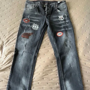 Svarta jeans med patchar och slitningar - Säljer ett par svarta jeans med coola patchar och slitningar framtill. Jeansen har raka ben och klassisk femficksdesign. Flera färgglada märken och detaljer ger ett unikt uttryck. Perfekta för dig som vill sticka ut lite extra.