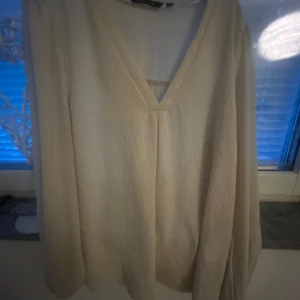 Beige blus med v-ringning från Amelie & Me - Säljer en stilren beige blus från Amelie & Me i storlek XS. Blusen har lång ärm och en djup v-ringning framtill. Perfekt för dig som gillar enkla och klassiska plagg med en avslappnad passform.