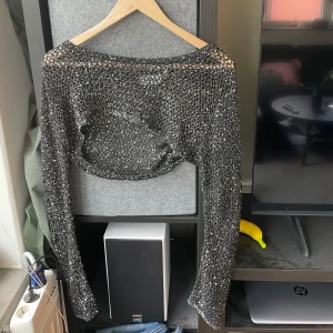 Glittrig croppad bolerotopp Weekday - Säljer en svart glittrig bolerotopp från Weekday, croppad modell med långa ärmar. Toppens yta är täckt av paljetter som ger en riktigt festlig look. Perfekt att styla över ett linne eller klänning för extra bling.