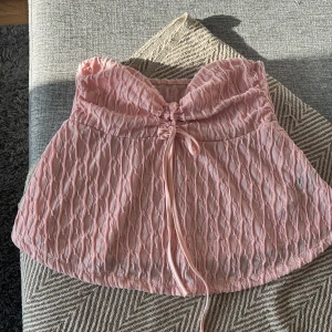 Rosa bandeau tubtopp med snörning - Storlek 38 ( passar 36) Supersöt rosa bandeau tubtopp med snygg struktur och knytband framtill. Toppens tyg har ett unikt mönster och lätt transparent känsla. Perfekt för varma dagar när du vill ha något luftigt och trendigt.