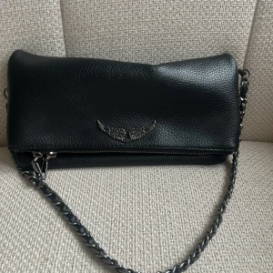 Zadig & Voltaire axelväska svart - Snygg svart axelväska i skinn med silverfärgad kedjerem och cool detalj med vingformad metall på framsidan. Väskan har dragkedja både fram och bak och en stilren, avlång form. Perfekt för dig som gillar edgy accessoarer med lite rockig känsla.Den är nästan oanvänd och ganska ren.Säljer då jag köpte en ny zadig väska som jag använder mest.