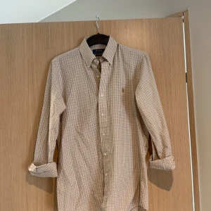 Beige-rutig skjorta Polo Ralph Lauren XS - Beige och vitrutig skjorta med lång ärm och broderad logotyp på bröstet. Skjortan har en button-down krage och manschetter.