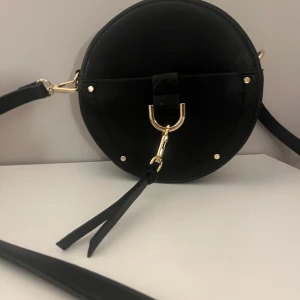 Rund svart axelväska med gulddetaljer - Säljer en rund svart axelväska i skinnimitation med guldfärgade metalldetaljer och dekorativ ring framtill. Väskan har en lång axelrem och en ficka på baksidan. Perfekt för att bära det viktigaste med stil.