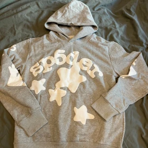 Grå hoodie med vita stjärnor från Sp5der - Säljer en grå hoodie från Sp5der med stora vita stjärnor och logga framtill samt tryck på ärmarna. Tröjan har huva och ribbade muddar vid ärmslut och nederkant. Perfekt för en avslappnad och trendig stil. Priset är ej hugget i sten.