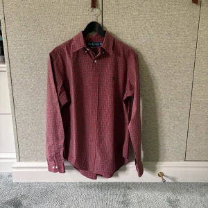 Rutig röd skjorta från Ralph Lauren - Säljer en klassisk röd och blå rutig skjorta från Ralph Lauren. Skjortan har lång ärm, knappar framtill och en liten broderad logga på bröstet. Perfekt för dig som gillar stilrena och tidlösa plagg. 
