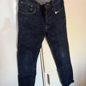 Nudie Jeans - Snygga mörkblå jeans från Nudie Jeans med klassisk femficksdesign och kontrasterande sömmar. Perfekta för en stilren look. De har en knappgylf och är tillverkade i slitstarkt denim.