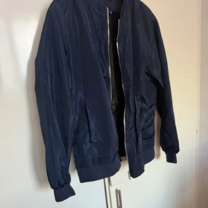 Mörkblå bomberjacka - Säljer en stilren mörkblå bomberjacka med dragkedja framtill. Jackan har långa ärmar och ribbade muddar vid ärmslut och krage. Perfekt för en casual look. Skulle säga den passar dig som har L- xl bättre då man kanske vill ha den lite oversize men det är min åsikt 