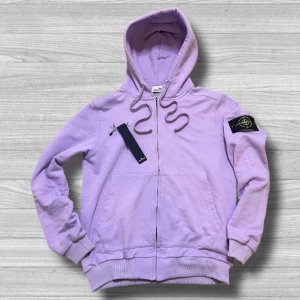 Stone Island - Säljer en snygg lila hoodie från Stone Island med dragkedja och huva. Tröjan har en ikonisk logga på ärmen och ribbade muddar vid ärmslut och nederkant. Perfekt för en avslappnad stil.
