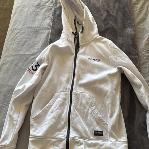 Vit huvtröja från Race Marine - Säljer en stilren vit huvtröja från Race Marine med dragkedja och tryck på ärmen. Tröjan har en justerbar huva med snörning och ribbade muddar vid ärmslut och nederkant. Perfekt för en avslappnad stil.