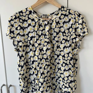 Blommig blus från Holly & White - Säljer en söt blommig blus från Holly & White i storlek xs. Blusen har ett härligt mönster med vita och gula blommor på en svart bakgrund. Den har korta ärmar och en rund halsringning med en liten knappdetalj i nacken. Inget tecken på användning🤗