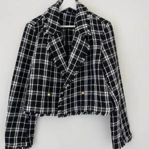 Svart och vit rutig kavaj - Snygg svart och vit rutig kavaj med fransiga detaljer och guldknappar. Den är croppad och har långa ärmar, perfekt för en stilren look. Passar bra till både vardag och festliga tillfällen.