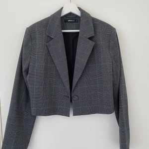 Grå rutig kavaj från Gina Tricot - Snygg grå rutig kavaj från Gina Tricot. Den är croppad med en klassisk krage och knäppning framtill. Perfekt för en stilren look.