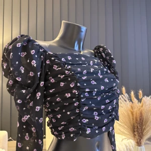 Svart blommig blus med puffärmar - Trendig svart blus med små lila blommor och puffiga långärmar. Blusen har en smickrande rynkad design och en elastisk rygg för extra komfort. Perfekt för att ge en romantisk touch till din outfit. Passar perfekt i S/M storlek.🌸👌