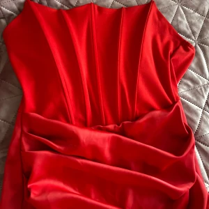 Röd långklänning -  röd långklänning med draperade detaljer framtill. Klänningen har en elegant och figurnära passform som framhäver silhuetten. Perfekt för speciella tillfällen.