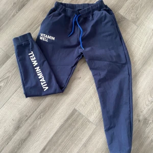 Mörkblå mjukisbyxor från Vitamin Well - Snygga mörkblå mjukisbyxor från Vitamin Well med vit logga på benet och vid fickan. Byxorna har en elastisk midja med blå snörning för justerbar passform. Perfekta för en avslappnad stil. Ambassadör collection går ej att köpa