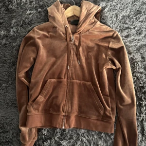 Brun zip up från Juicy Couture - Säljer en brun hoodie från Juicy Couture i mjukt material med dragkedja och huva. Den har en snygg logga på bröstet och praktiska fickor framtill. Tecken på användning men överlag bra skick. Säljer settet för 500kr, annars byxorna för sig 300 och tröjan för sig 300.