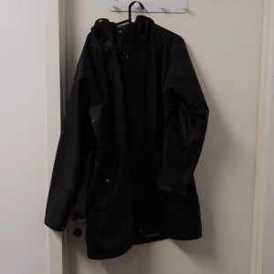 Svart regnjacka från Cross Sportswear - Säljer en stilren svart regnjacka från Cross Sportswear. Jackan har långa ärmar och en praktisk huva. Perfekt för regniga dagar och håller dig torr och bekväm. Den har en enkel design med diskreta detaljer.