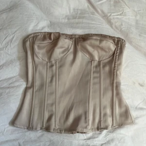 Beige korsettopp med dragkedja - Snygg beige korsettopp med dragkedja bak. Toppen är axelbandslös och har en stilren design med markerade sömmar. Perfekt för att skapa en elegant look.
