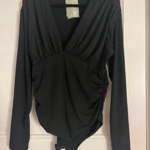 Svart långärmad omlottopp - Snygg svart body med långa ärmar och djup v-ringning från H&M. Toppen har rynkade detaljer på sidorna för en smickrande passform. Perfekt för en stilren look. Aldrig använd och säljer den för att den inte är rätt i storlek!