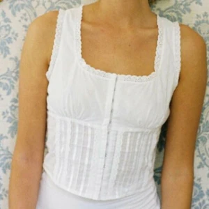 With Jean Adeline Top (Vit korsettopp med spetsdetaljer) - Säljer en vit korsett med spetsdetaljer och knäppning fram. Den har en snygg snörning bak och är ärmlös. Perfekt för att ge en stilren look.
