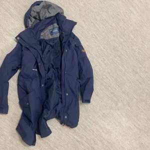 Blå parkajacka från Tenson - Säljer en snygg blå parkajacka från Tenson. Jackan har en praktisk huva och flera fickor med knappar. Perfekt för kyligare dagar med sin långa design och dragkedja framtill. Märkesdetaljer på ärmen ger en extra touch.