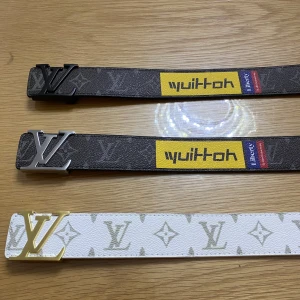 Bälten från Louis Vuitton - Tre stiliga bälten från Louis Vuitton. Två i brunt med silverfärgat spänne och ett i vitt med guldfärgat spänne. De bruna bältena har en gul och röd detalj med text, medan det vita bältet har ett klassiskt monogrammönster.