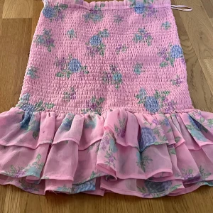 Rosa blommig kjol från BikBok stl xs - Charmig rosa kjol från BikBok med blommigt mönster i blått och grönt. Kjolen har en volangdetalj längst ner och är elastisk för en bekväm passform. Perfekt för en somrig look!