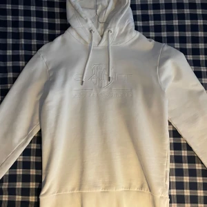 Vit hoodie från GANT - Säljer en stilren vit hoodie från GANT med broderad logga på bröstet. Hoodien har en justerbar dragsko i huvan och ribbade muddar vid ärmslut och nederkant. Perfekt för en avslappnad stil. Inga skador finns på denna. Skulle säga att den är lite liten i storleken.
