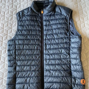 Save the duck väst  - Snygg blå dunväst från Save The Duck med dragkedja framtill. Västen är quiltad och tillverkad i syntetiskt material, Plumtech, som är både vattenavvisande och andas. Perfekt för kyligare dagar. Liten i storleken. 