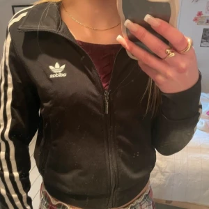 Svart träningsjacka från Adidas - Säljer en klassisk svart träningsjacka från Adidas med de ikoniska tre vita ränderna längs ärmarna. Jackan har dragkedja framtill och två sidofickor med dragkedjor. Har en liten fläck men inget som man tänker på. Xxs 