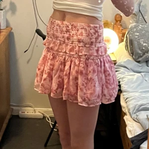 Rosa blommig kjol - Söt rosa kortkjol med blommigt mönster och volangdetaljer. Från Gina och är nedsydd i midjan med några stygn som enkelt går att sprätta upp om så önskas. Använd fåtal gånger. Jag är 172cm