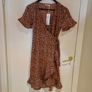 Brun blommig omlottklänning från ONLY stl 36 - Charmig brun omlottklänning med blommigt mönster. Klänningen har korta ärmar och volangdetaljer vid fållen. Perfekt för en avslappnad och stilfull look.
