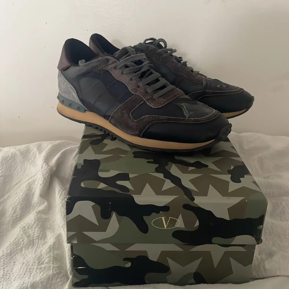 Snygga Valentino Rockrunner sneakers med ett coolt camouflagemönster. Skorna har en kombination av skinn och mocka i olika nyanser av svart och grått. De har snörning och en bekväm sula i beige. Perfekta för en stilren look. En äldre model. Kengät.
