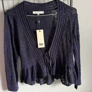 Mörkblå spetskofta från Flash Woman - Säljer en elegant mörkblå spetskofta från Flash Woman. Koftan har långa ärmar och en v-ringad design med knytband framtill. Den har ett vackert spetsmönster och volangdetaljer vid nederkanten. 🦋