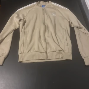 Beige jacka/zip från Adidas - Säljer en beige jacka/tröja från Adidas med dragkedja och de klassiska tre ränderna längs ärmarna. Fungerar för både träning och vardag.