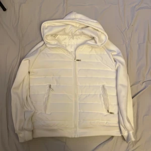 Vit cardigan  - Säljer en stilren vit hoodie med quiltad design och dragkedja. Den har en praktisk huva och två fickor med dragkedjor. Perfekt för en avslappnad look.