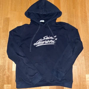 Svart hoodie från Saint Laurent - Snygg svart hoodie från Saint Laurent med logga i vitt på bröstet. Den har en klassisk känguruficka och justerbar huva med snören. Perfekt för en stilren och bekväm look. Pris går att diskutera. 
