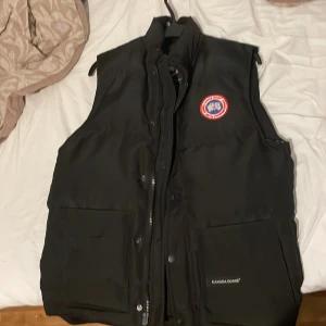 Svart dunväst från Canada Goose - Säljer en svart dunväst från Canada Goose med en klassisk design. Västen har en broderad logotyp på bröstet och praktiska fickor framtill. Perfekt för kyliga dagar.